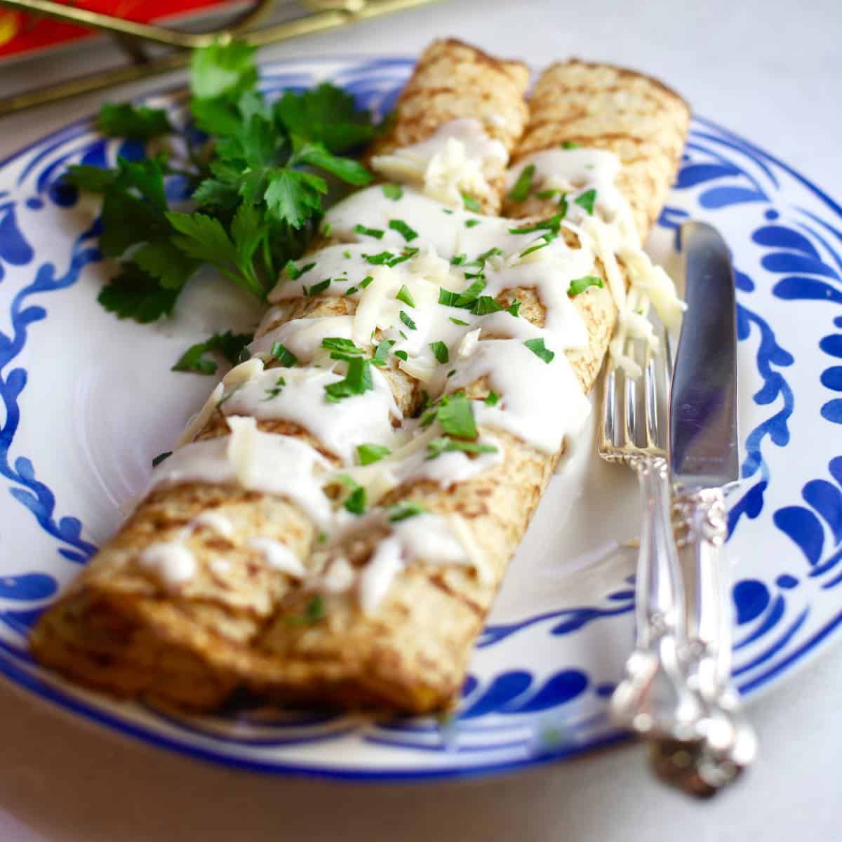 Two chicken and mushroom crepes topped with mornay sauce on a blue and white plate.