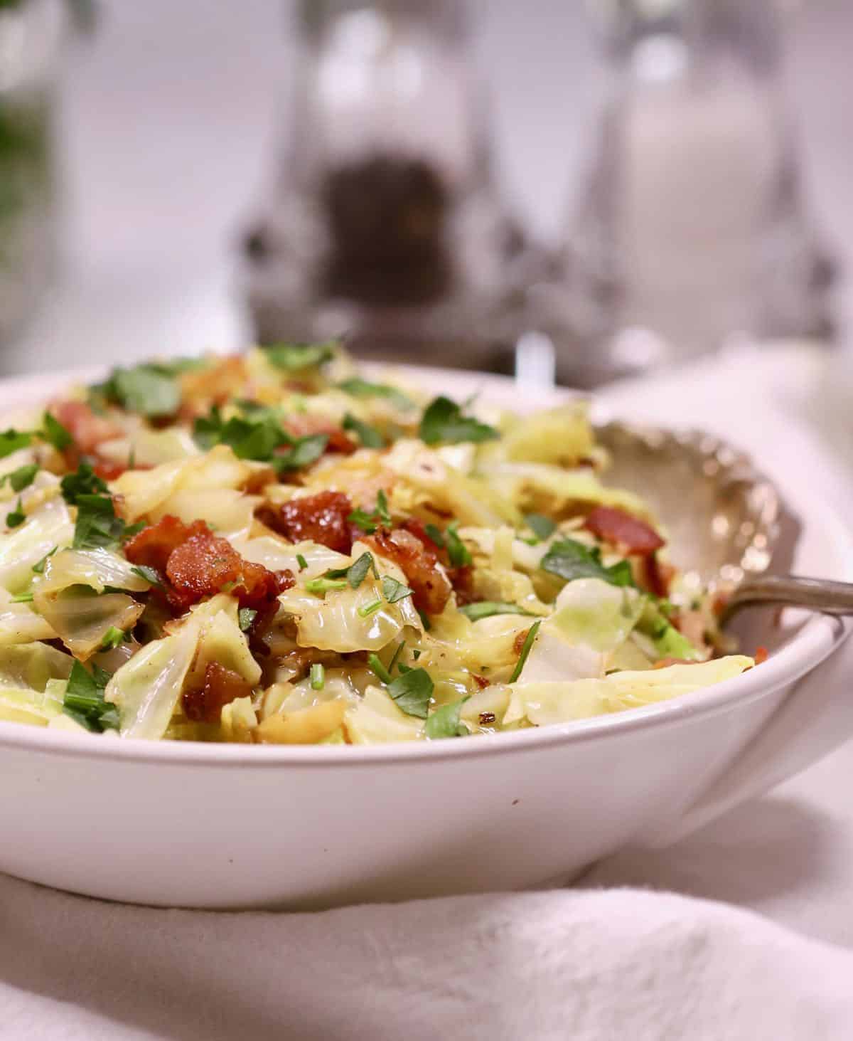 A white serving bowl full of fried cabbage topped with bacon. 
