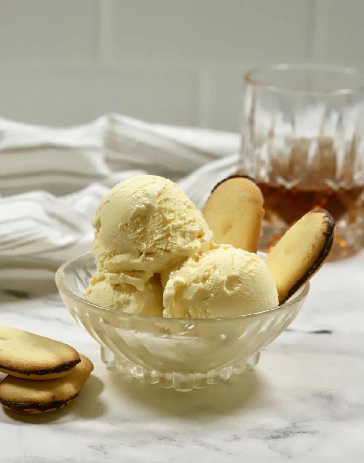 Three scoops of bourbon ice cream in a glass bowl with a glass of bourbon in back.