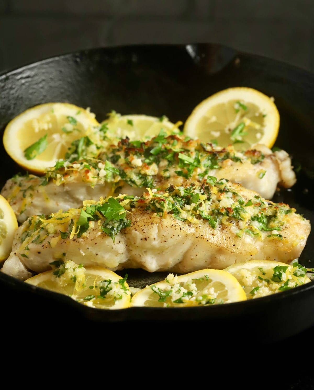 Two grouper fillets topped with gremolata in a cast-iron skillet with lemon slices. 