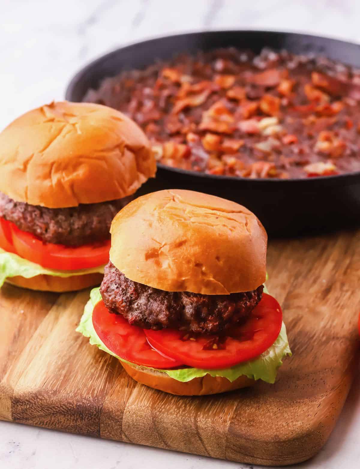 Two burgers on a wooden cutting board with a pan of baked beans.