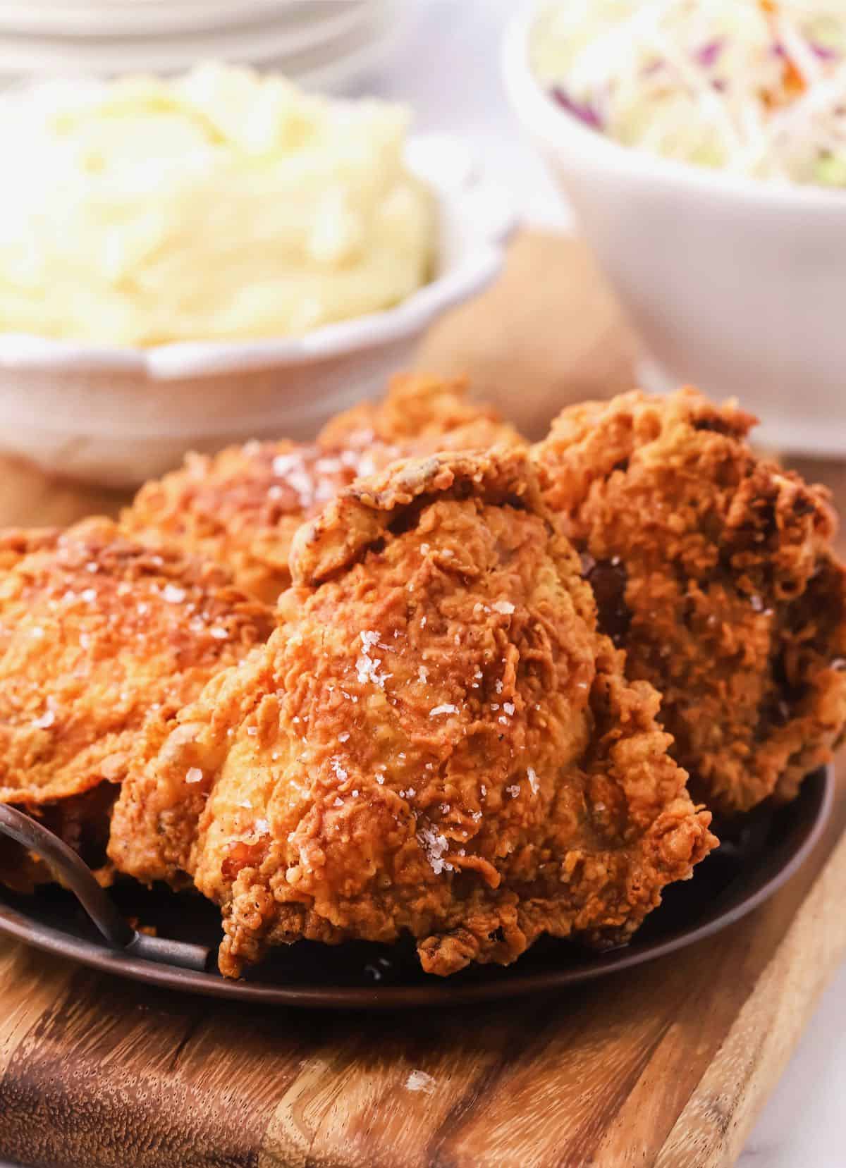 Fried chicken on a plate with a bowl of mashed potatoes in the background. 