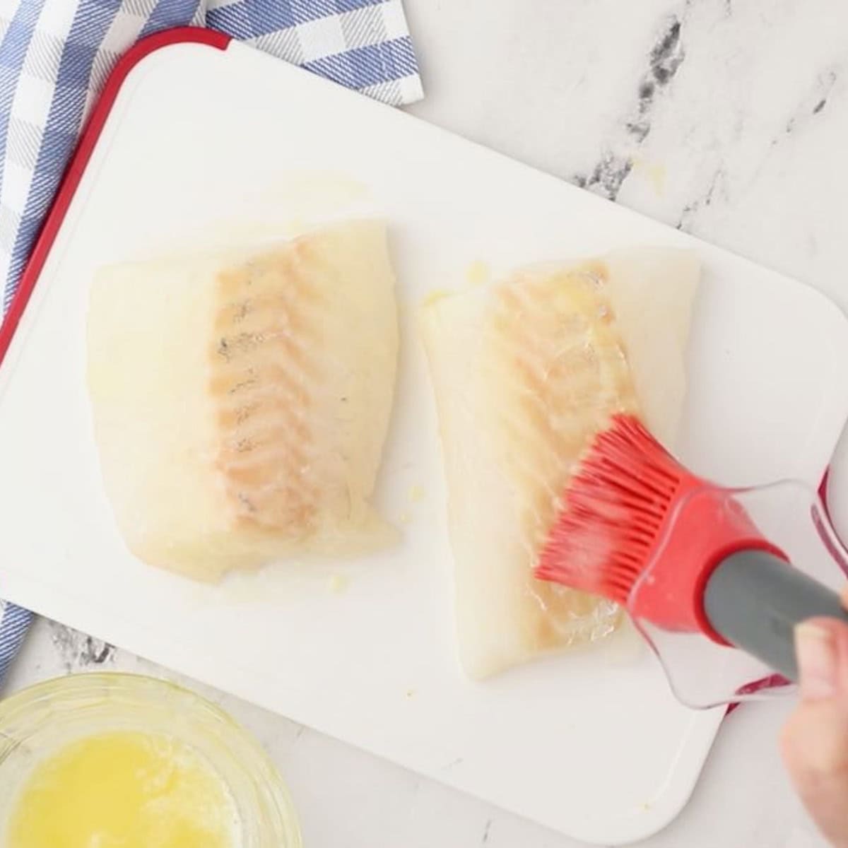 Using a pastry brush to brush melted butter on two cod fillets.