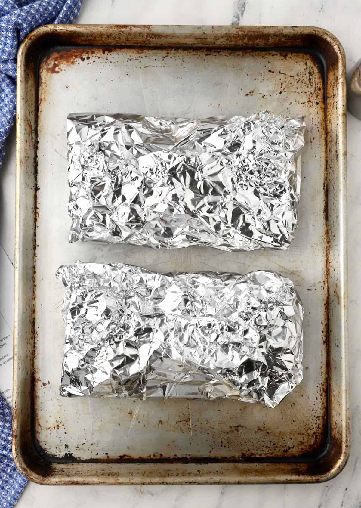 Two racks of ribs wrapped in aluminum foil on a baking sheet.