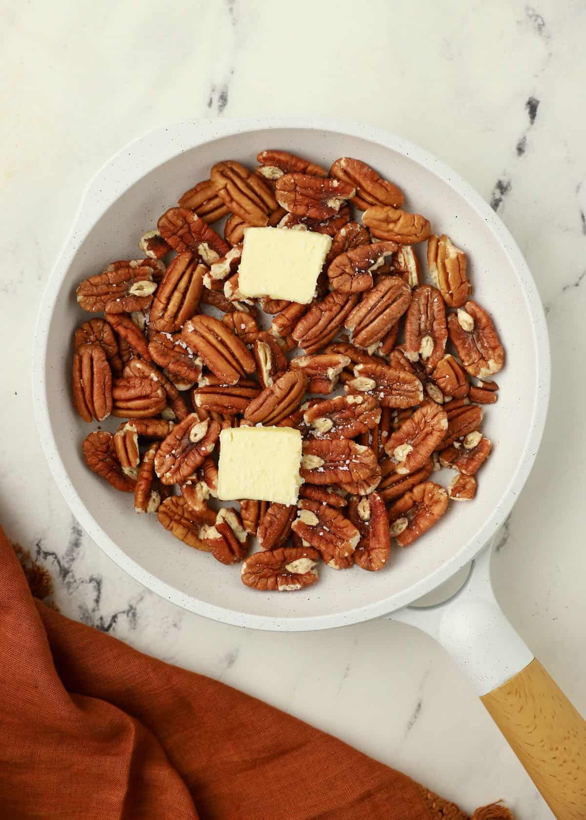 A white skillet full of pecan halves topped with two pats of butter. 