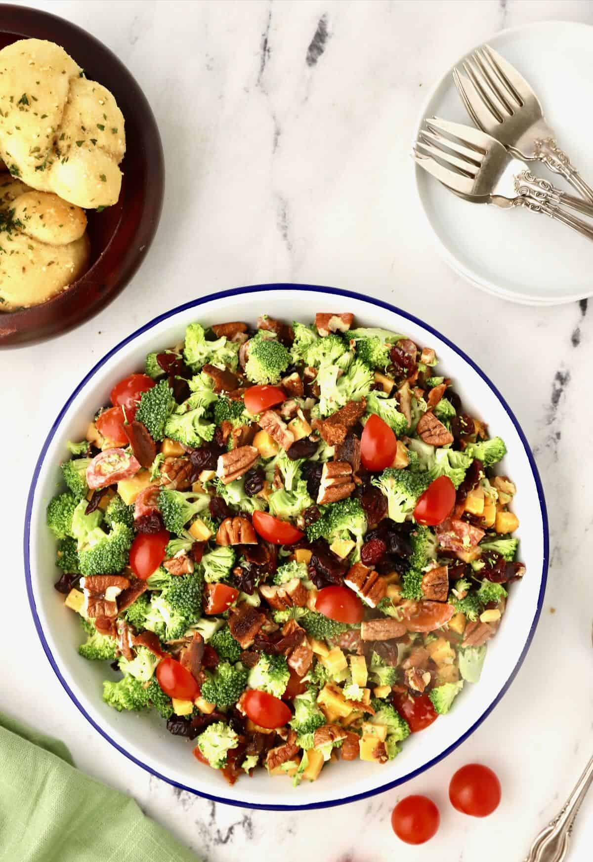 Broccoli Salad in a white metal bowl with rolls and plates. 