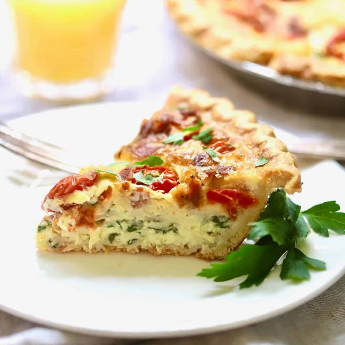 A slice of spinach and bacon quiche with tomatoes on a white plate.