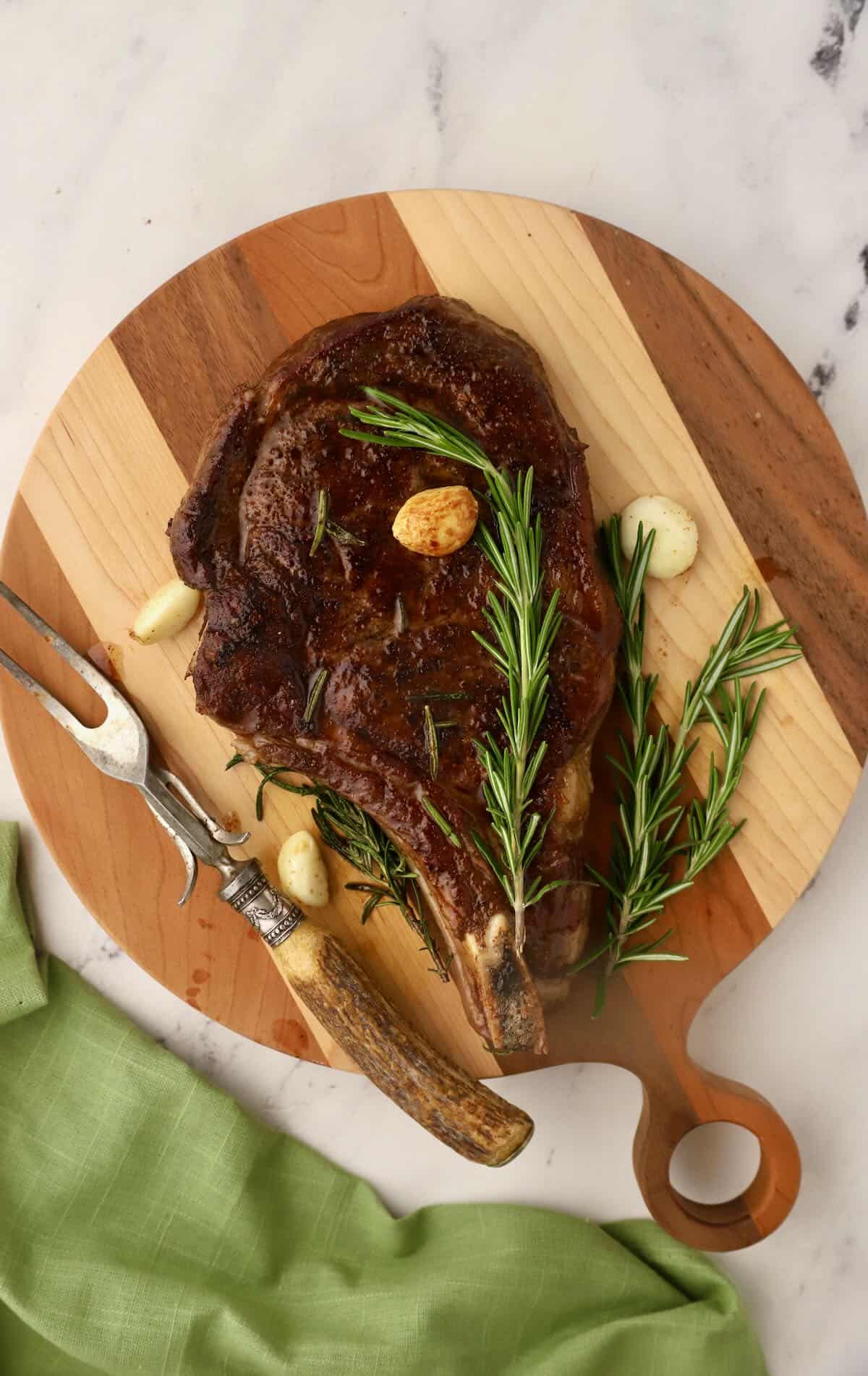 A steak on a cutting board topped with garlic cloves and rosemary. 