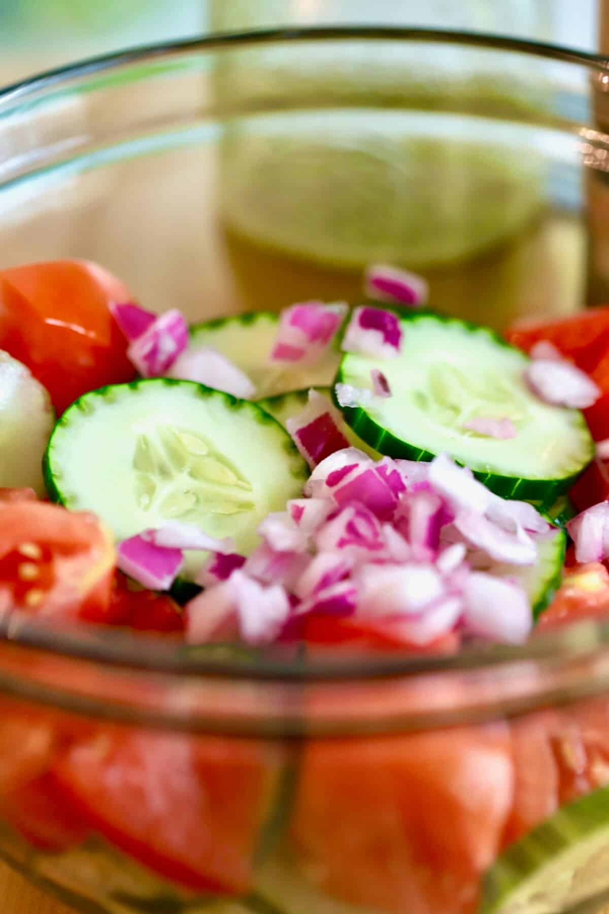 Sliced cucumbers, tomatoes and diced red onion in a glass bowl.