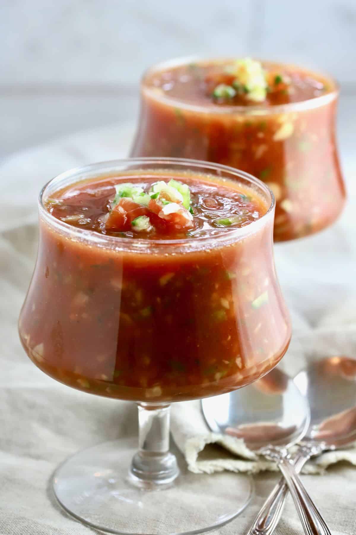 Two glass dishes full of gazpacho with spoons. 
