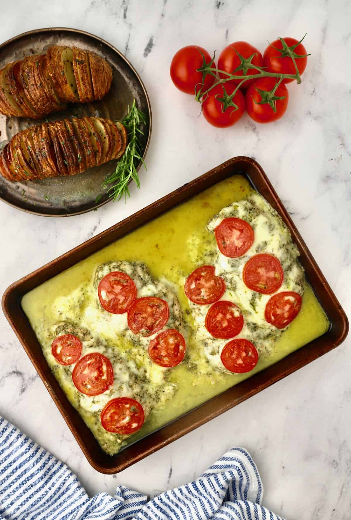 Baked chicken breasts topped with pesto, cheese and tomatoes on a baking sheet. 