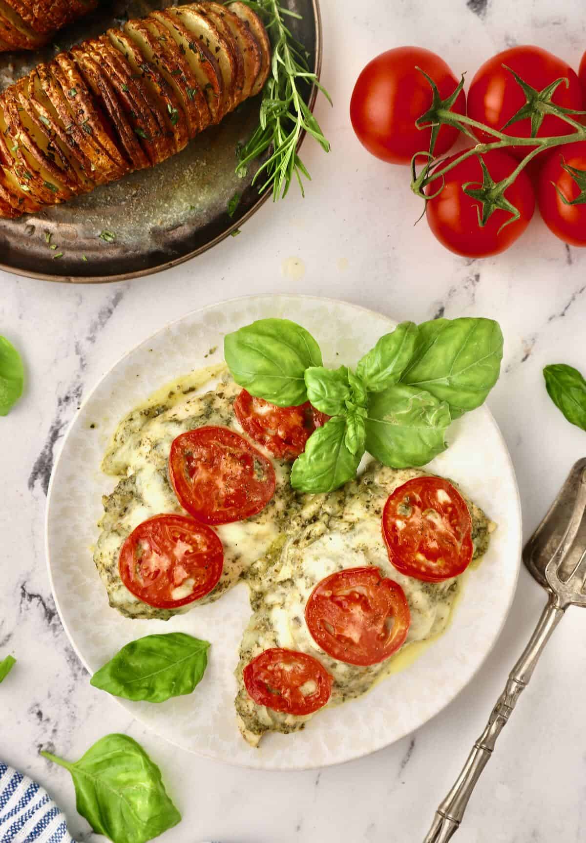 Two baked chicken breasts on a plate topped with pesto, cheese, and tomatoes. 