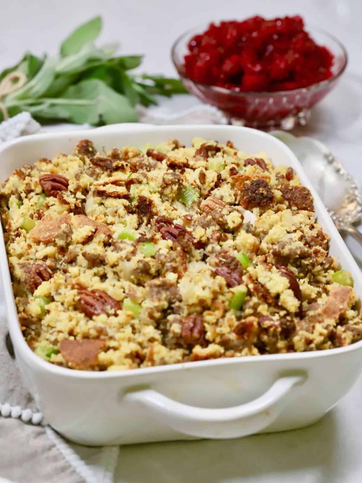 Baked cornbread dressing with sausage and pecans in a baking dish with cranberry sauce behind it.