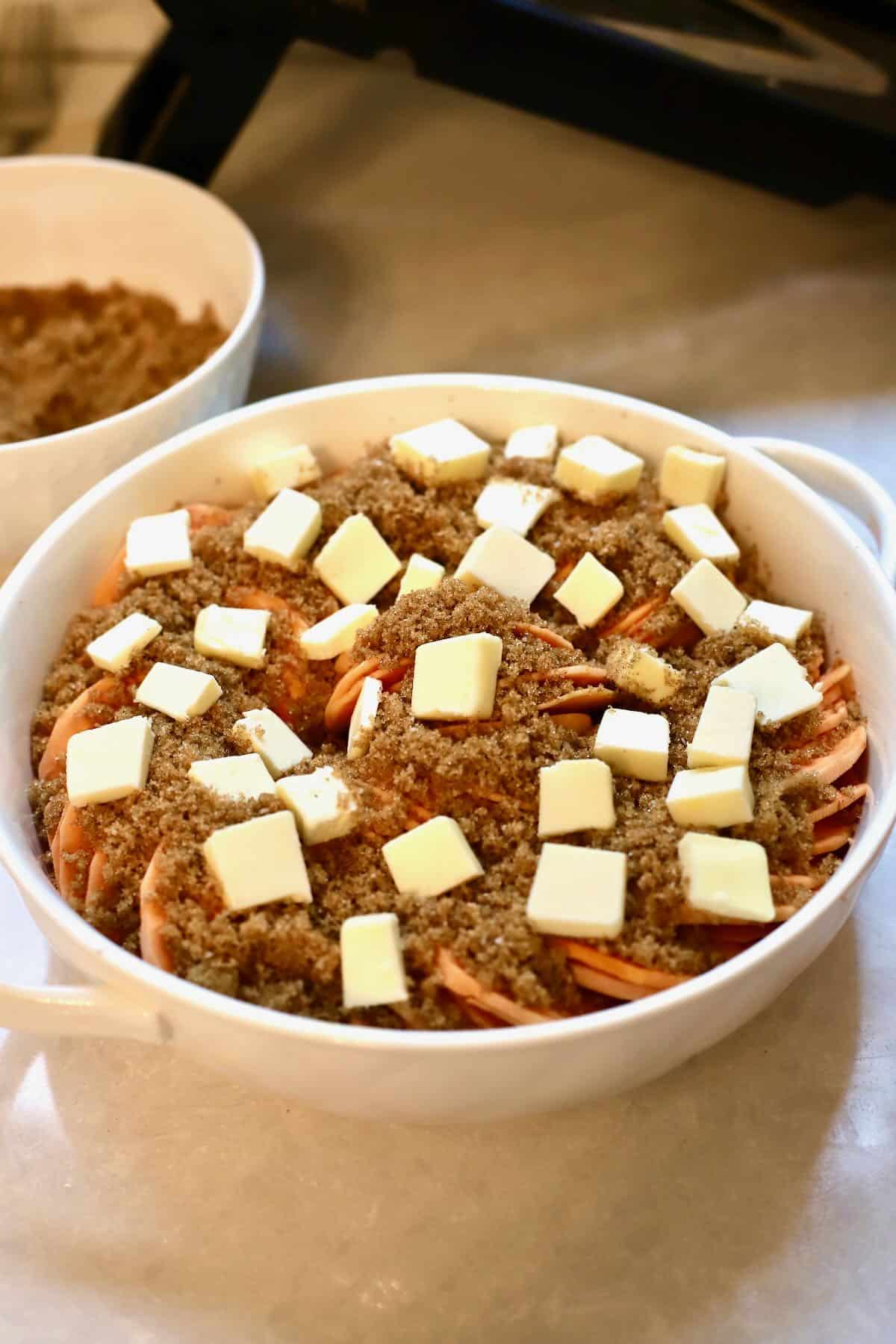 A casserole dish with sliced potatoes topped with brown sugar and cubes of butter. 