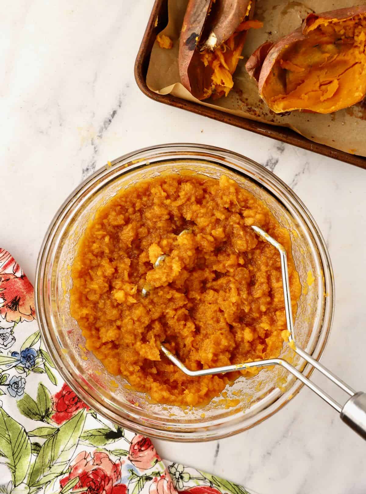 A clear glass bowl with sweet potatoes being mashed with a potato masher. 