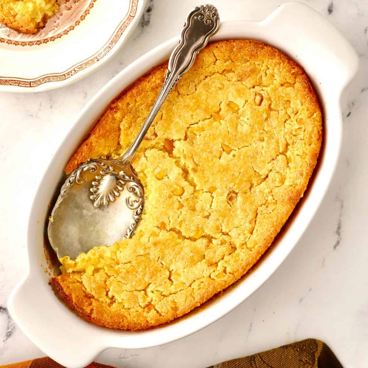 An oval casserole dish with cornbread pudding and a serving spoon.