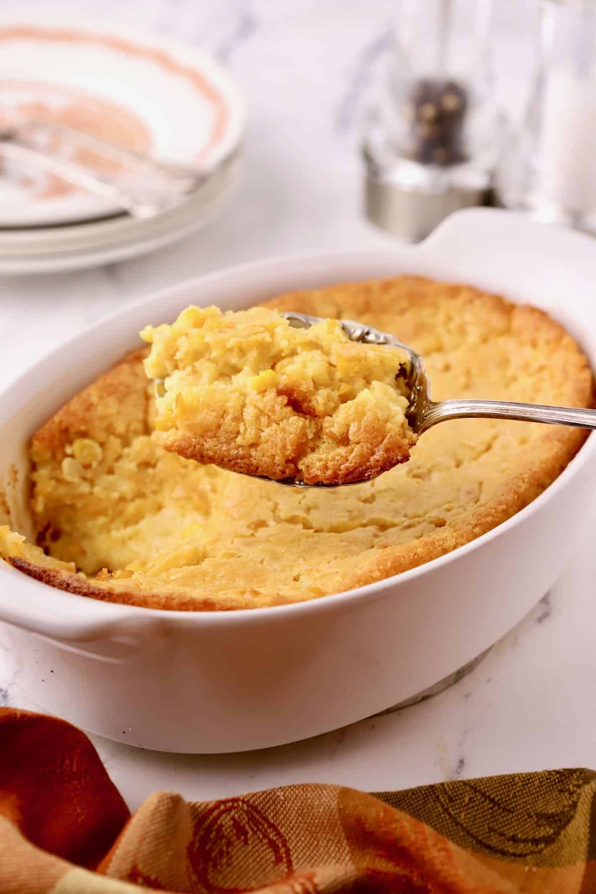 A white casserole dish full of cornbread pudding and a serving spoon with a serving of it. 