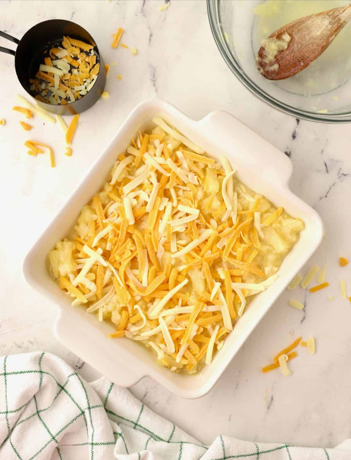 Square white baking dish with casserole ingredients topped with shredded cheese. 