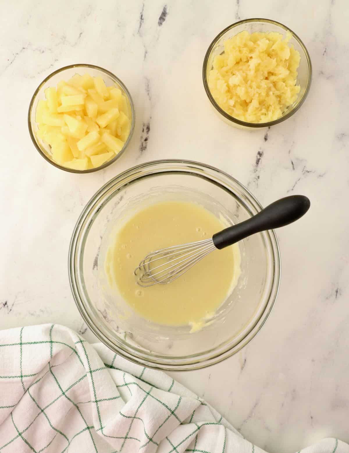 A clear glass bowl and whisk with flour, sugar and pineapple juice mixture. 