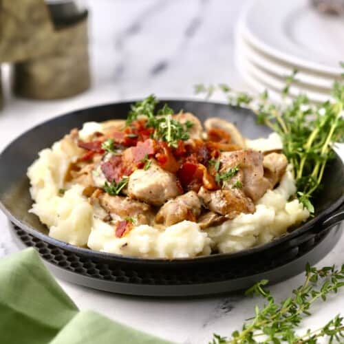 A small skillet with mashed potatoes topped with chicken, mushrooms and bacon.