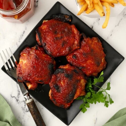 Four BBQ chicken thighs on a square black plate.