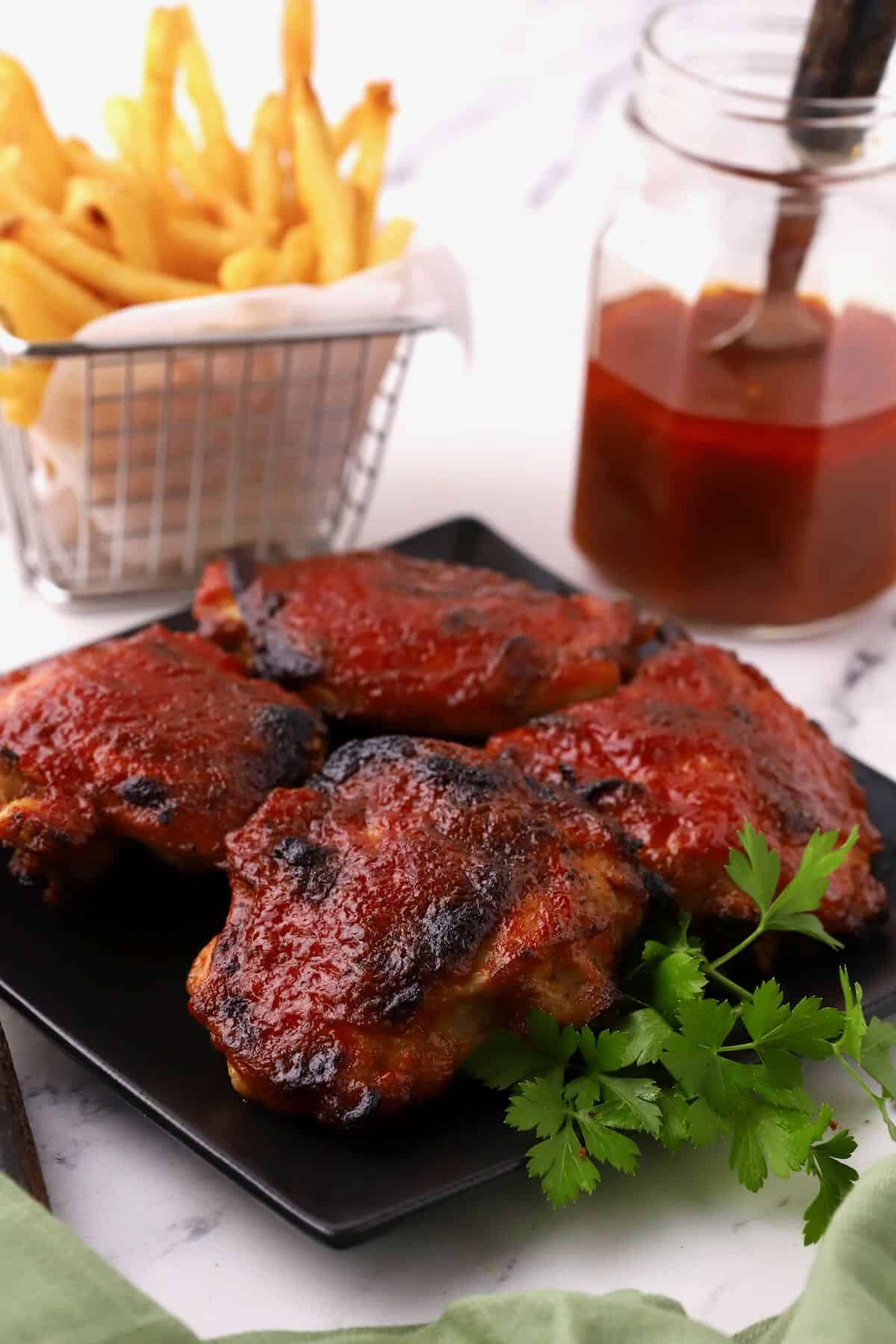 BBQ chicken thighs on a black plate with a basket of fries in the background. 