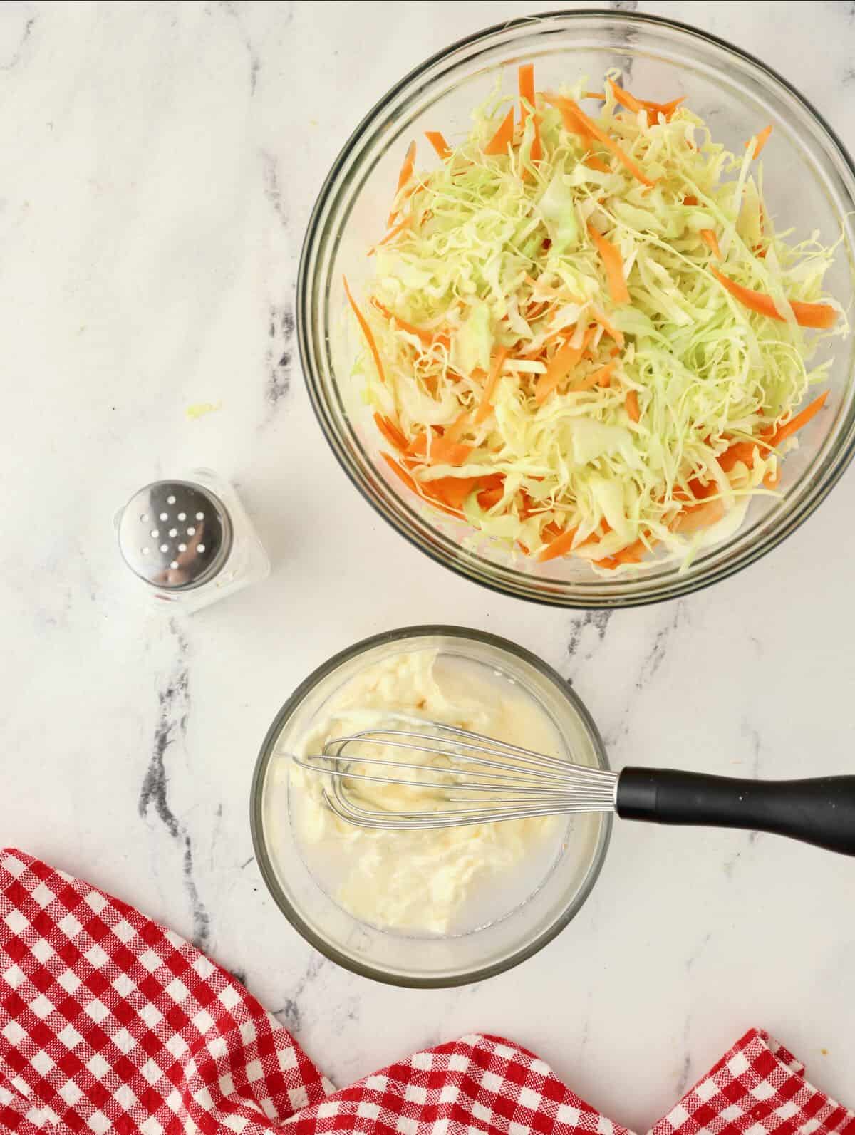A bowl of coleslaw and a bowl of dressing with a whisk. 