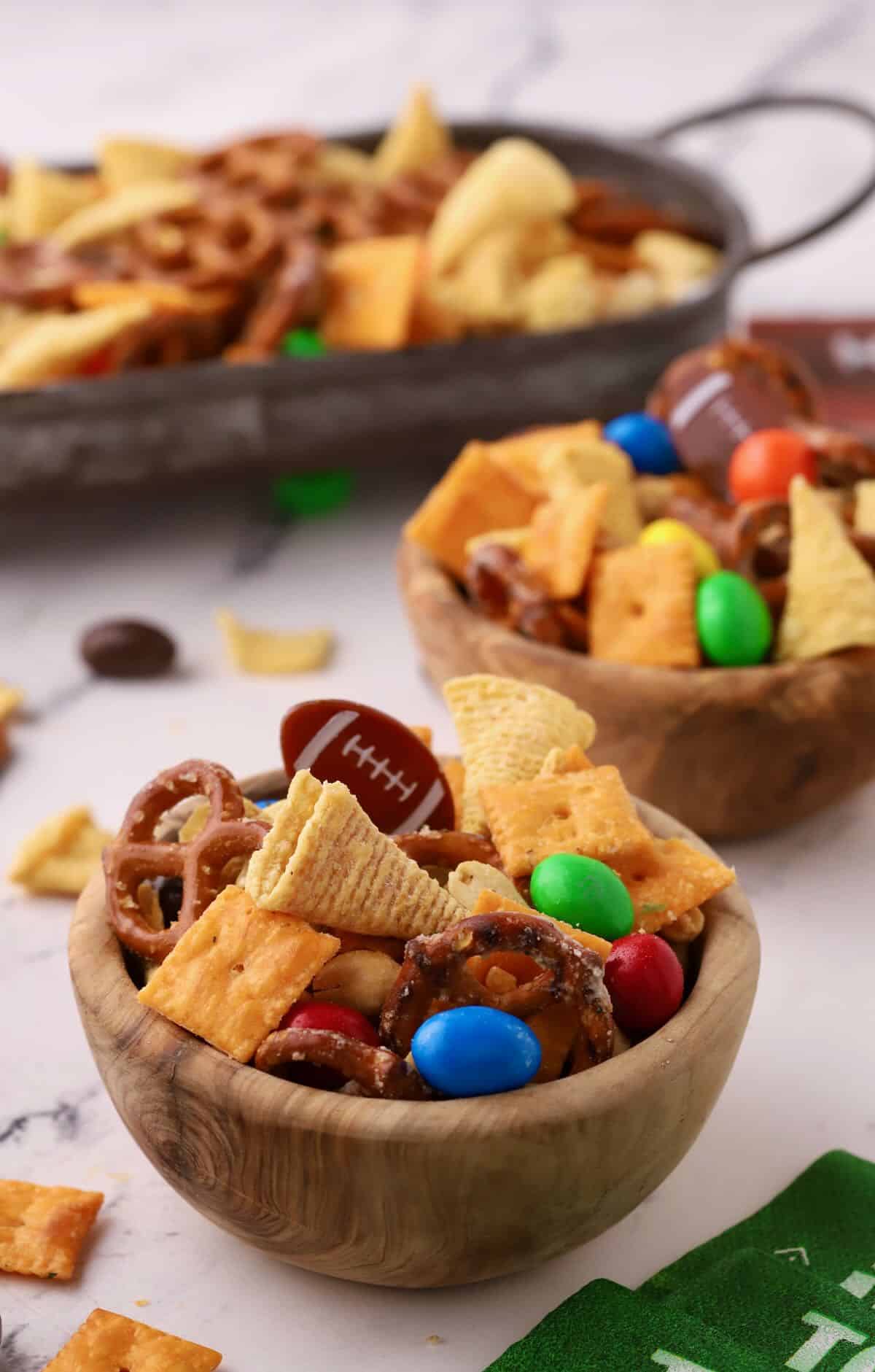Two small wooden bowls of a snack mix containing M&Ms, Cheeze-its, and Bugles. 