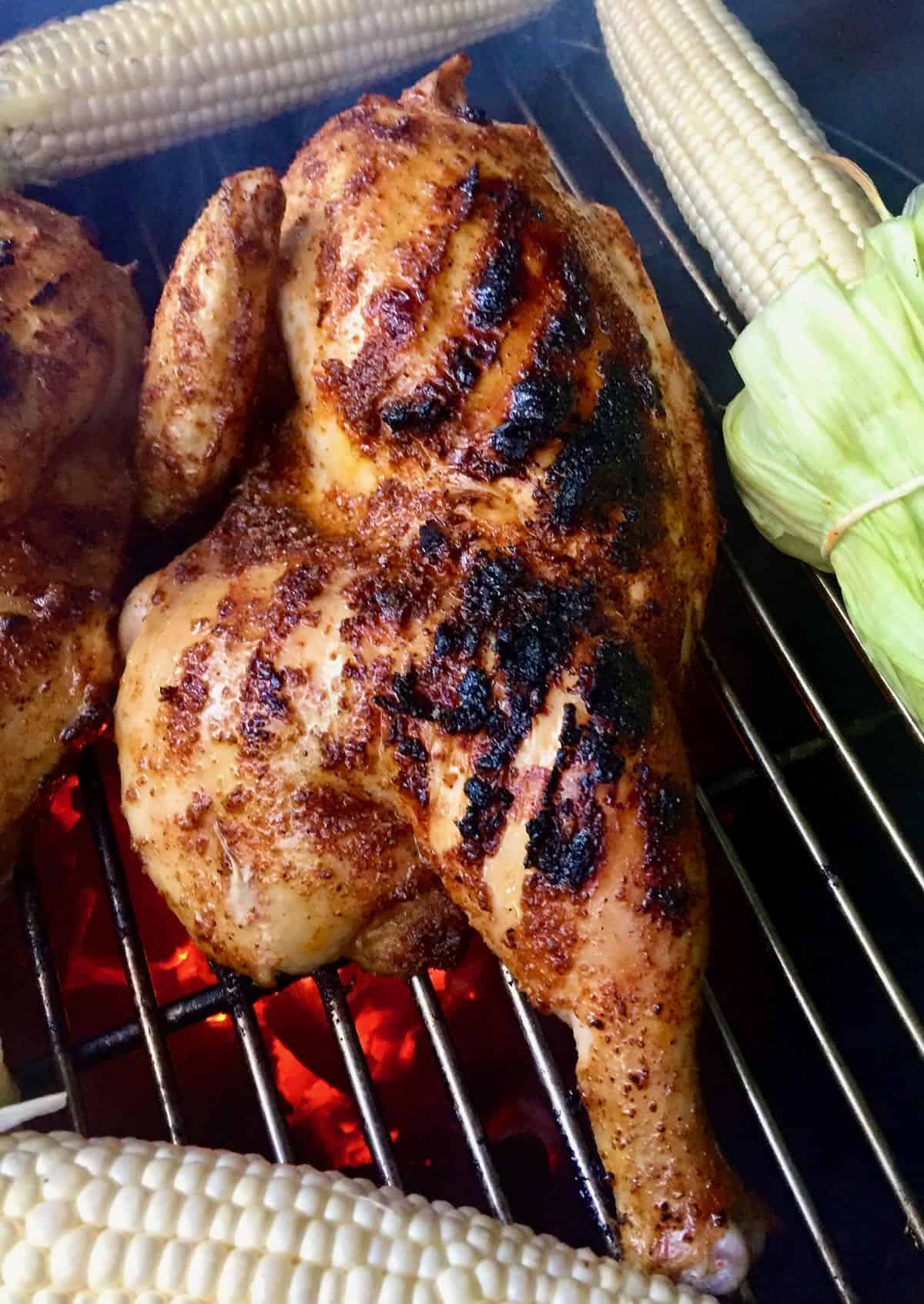 Grilled half chicken surrounded with sweet corn cooking on grill grates. 