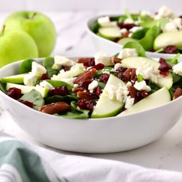 Spinach salad topped with sliced apples, pecans and feta cheese in a white bowl.