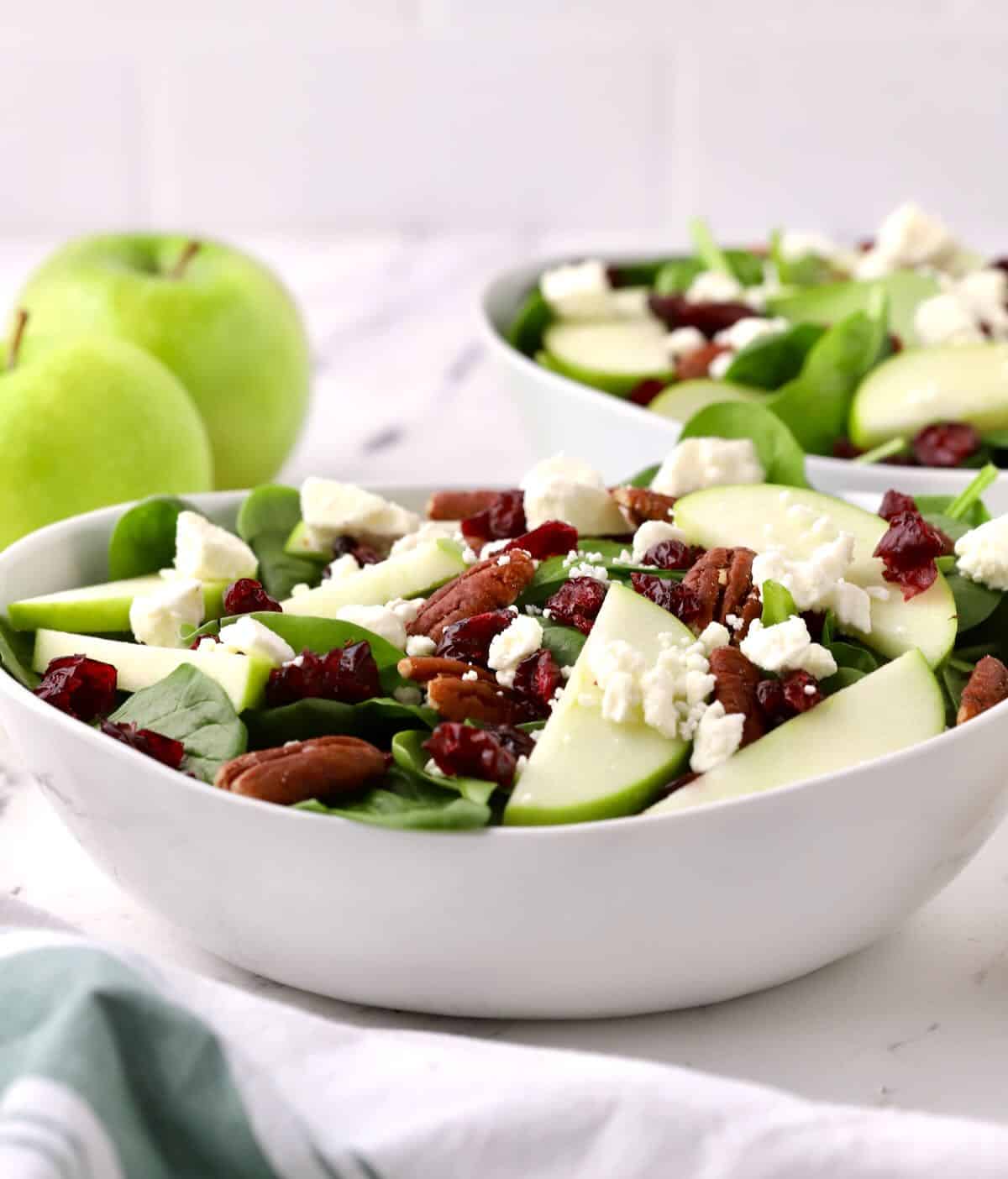 Spinach salad topped with apples, pecans and feta in a white bowl. 