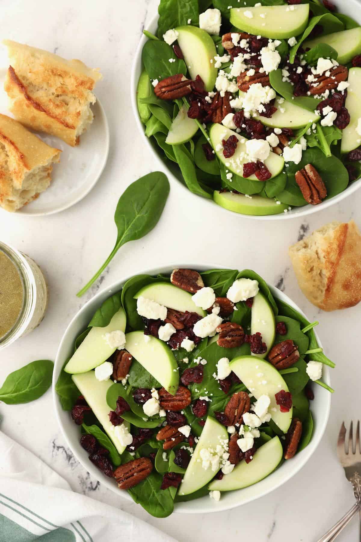 Two bowls full of spinach salad topped with apples and pecans. 