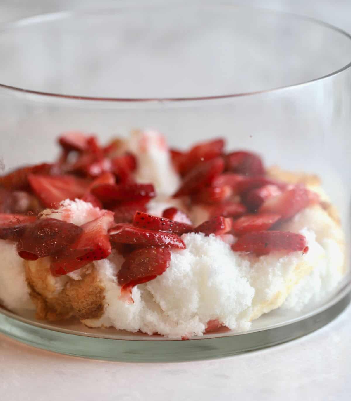 A layer of pieces of angel food cake and sliced strawberries in a glass trifle dish. 