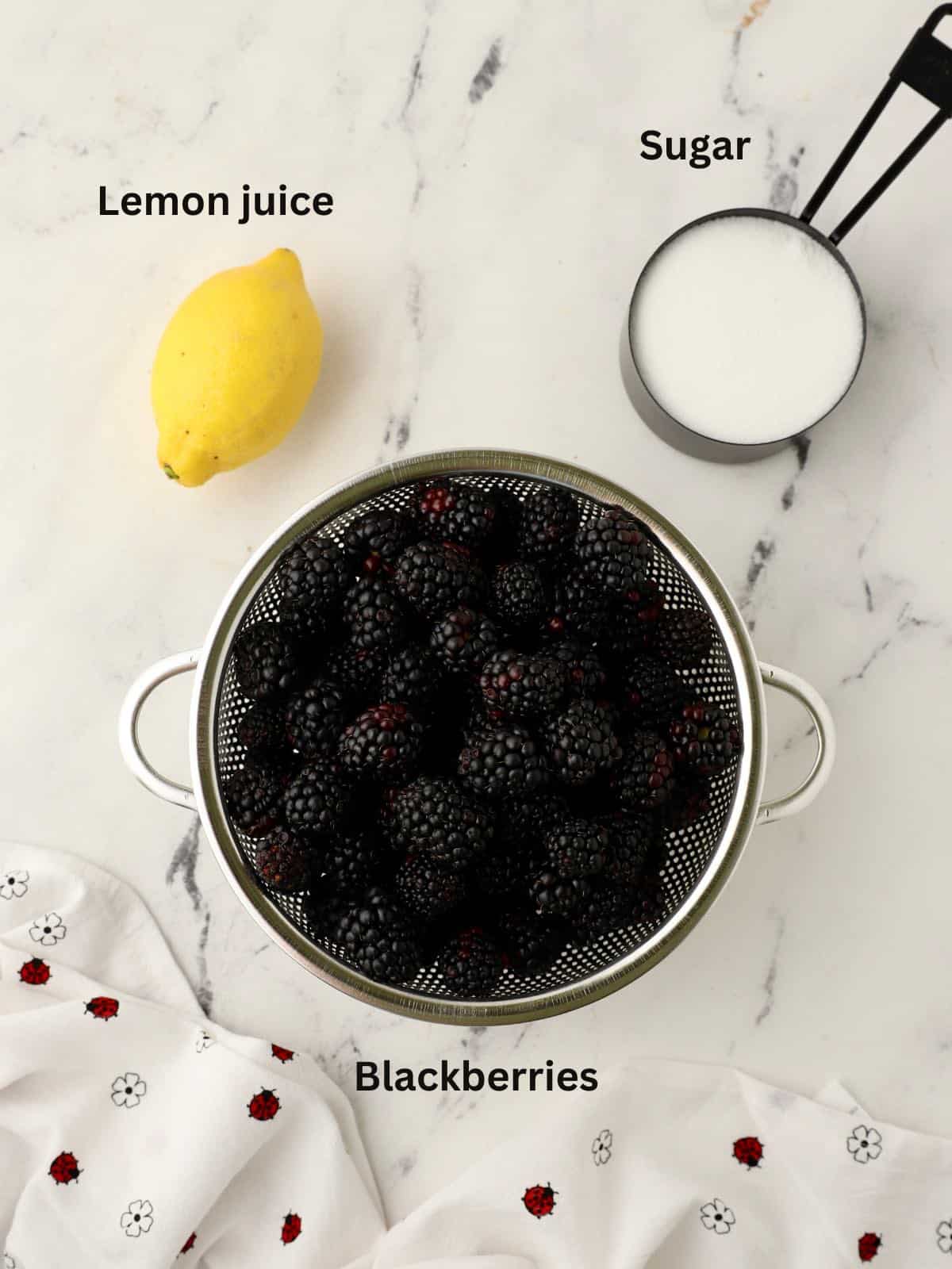 A colander full of blackberries, a measuring cup of sugar and a lemon.