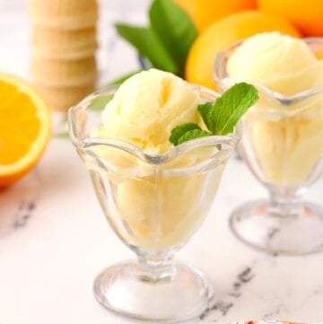 A clear glass dish with two scoops of orange sherbet, garnished with a sprig of mint.