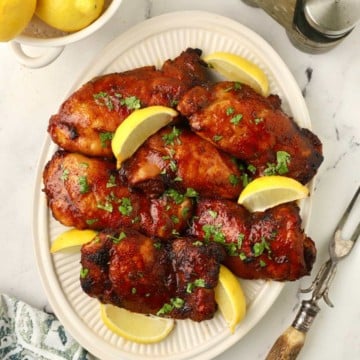 Broiled chicken thighs on a white plate, garnished with lemon slices.