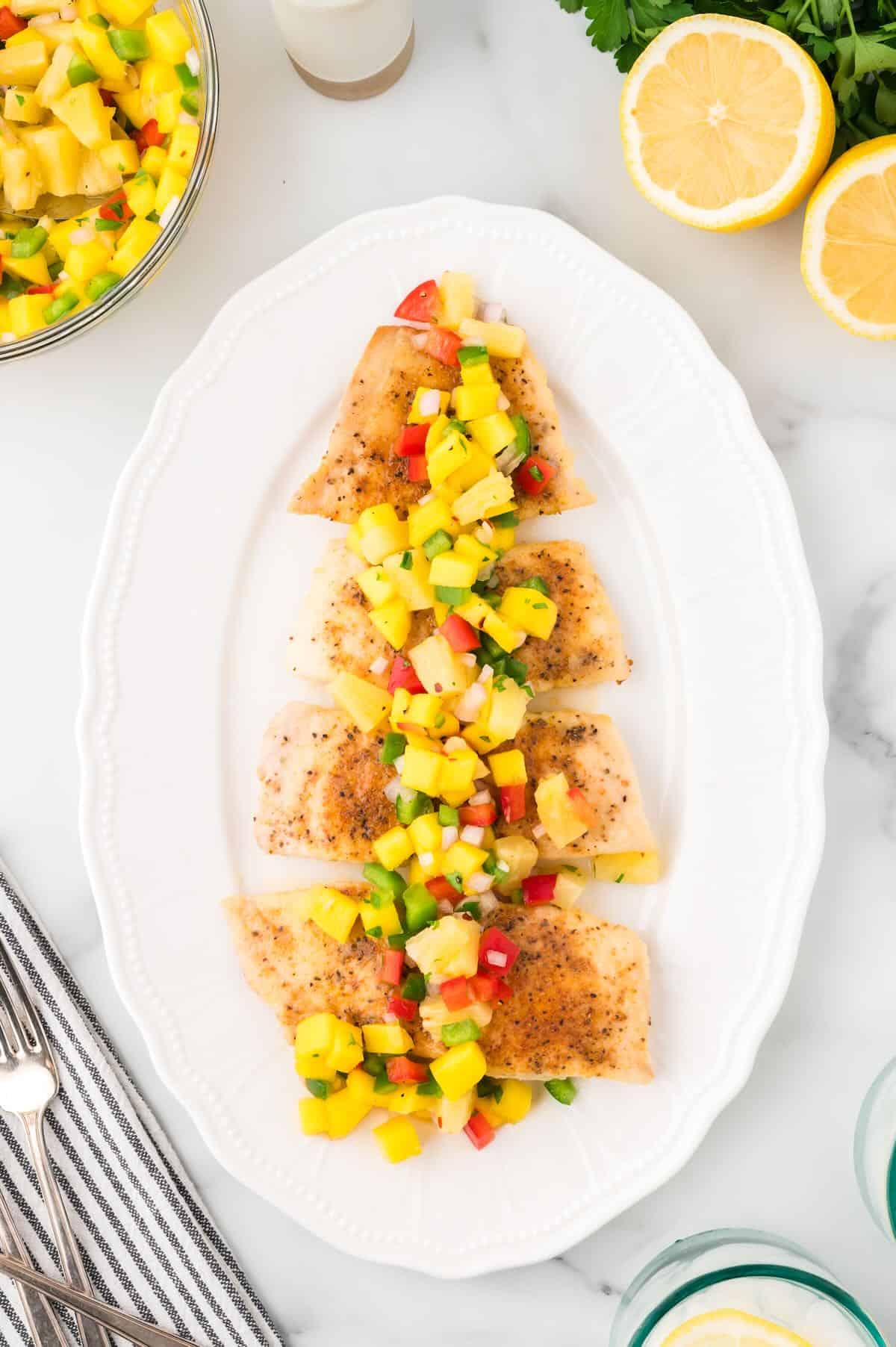 A white serving platter with pan-seared snapper fillets cut into pieces and topped with mango salsa.