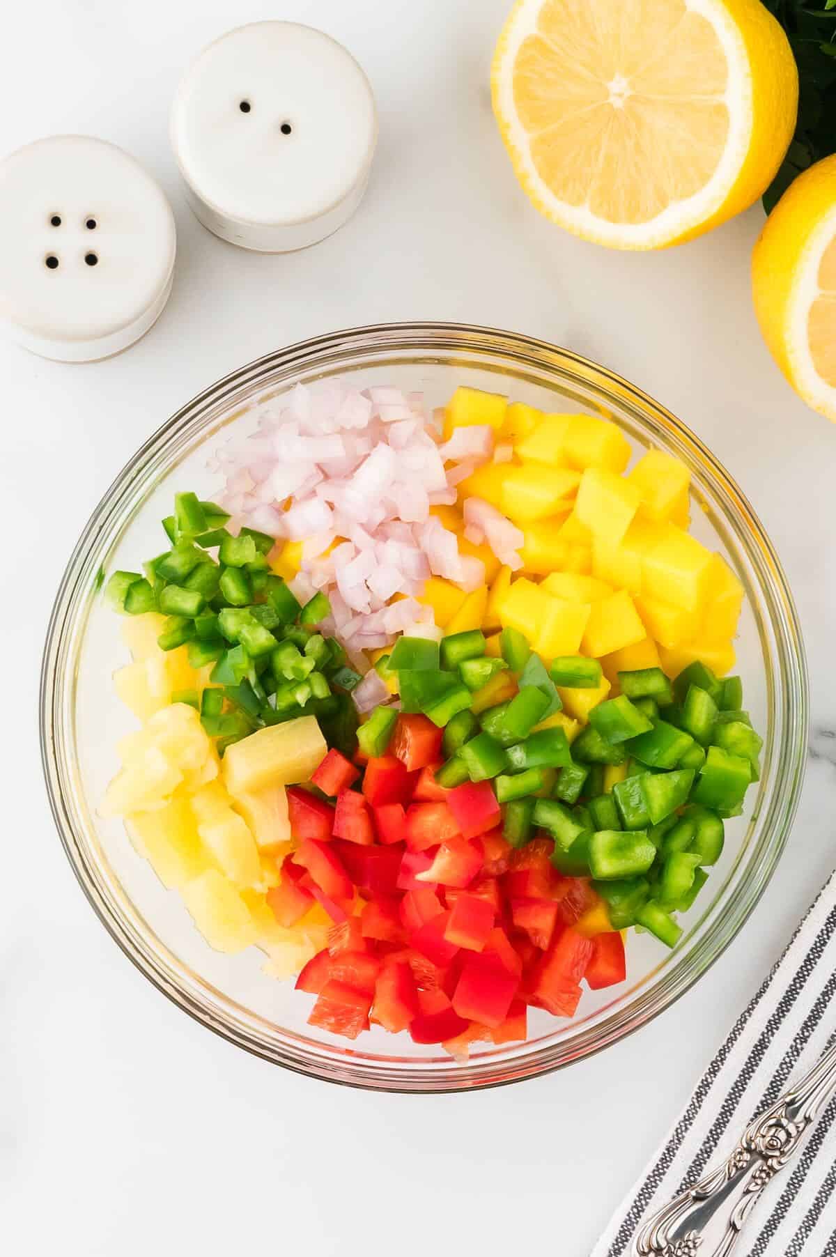A large clear glass bowl full of chopped mango, bell peppers, pineapple, and jalapeno. 