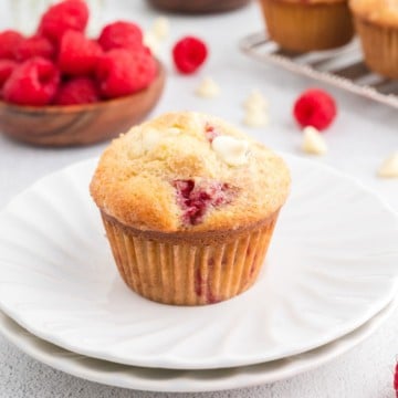 A raspberry white chocolate muffin in a white plate with a bowl of raspberries in the background.