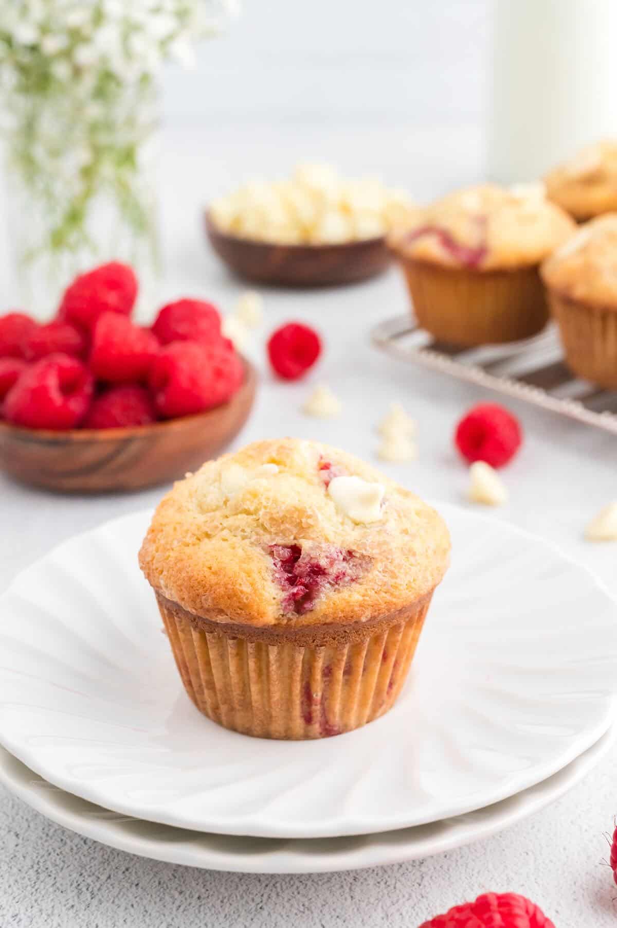 A raspberry white chocolate chip muffin on a stack of white fluted plates. A silver cooling rack with more muffins is in the background, along with wooden dishes of raspberries and white chocolate chips.