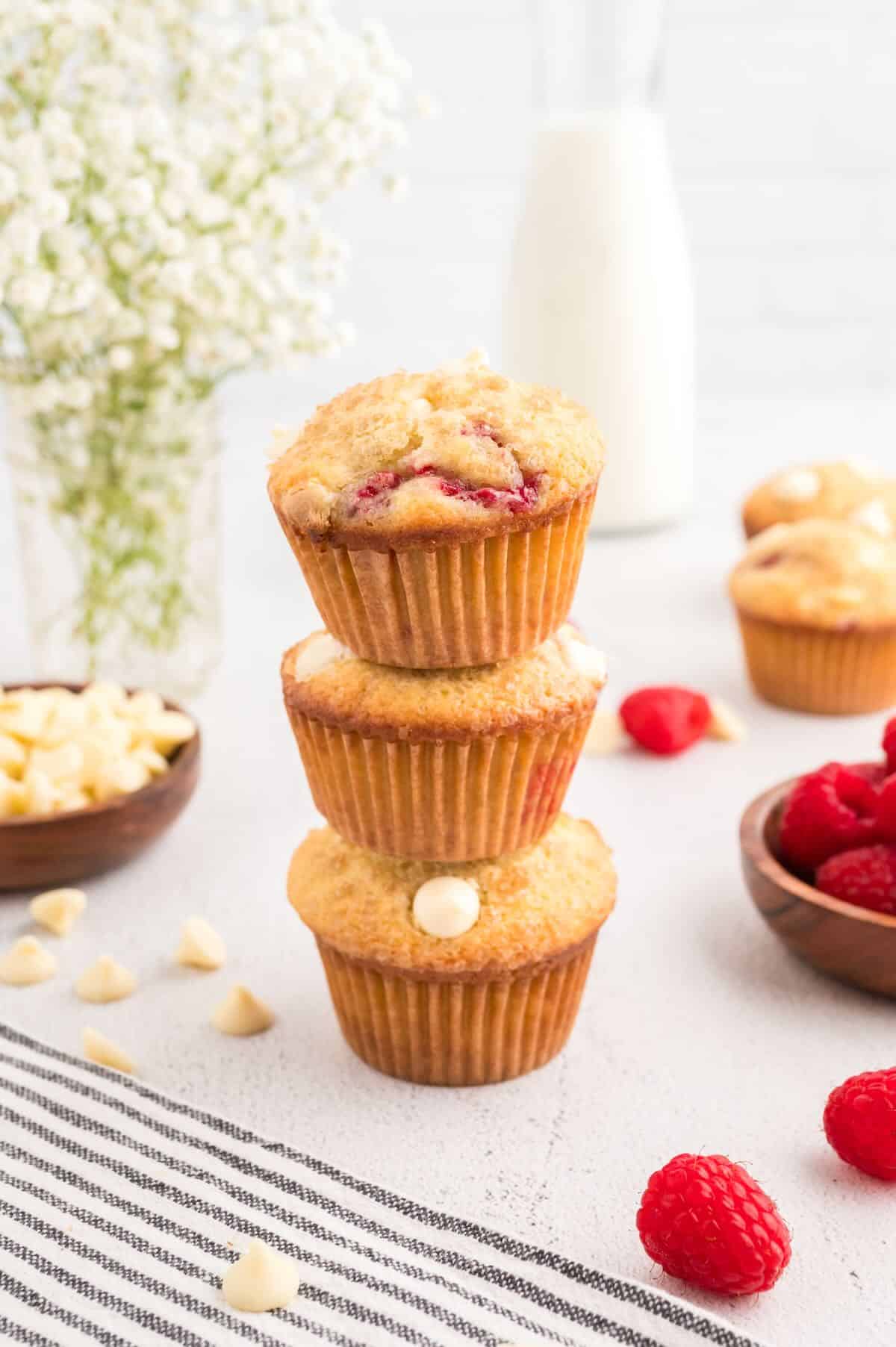 A stack of three raspberry white chocolate muffins on a white background. A jug of milk and vase of white flowers are the in the background. More muffins, and dishes of white chocolate and raspberries are to the side. Raspberries and white chocolate pieces are scattered across the background.