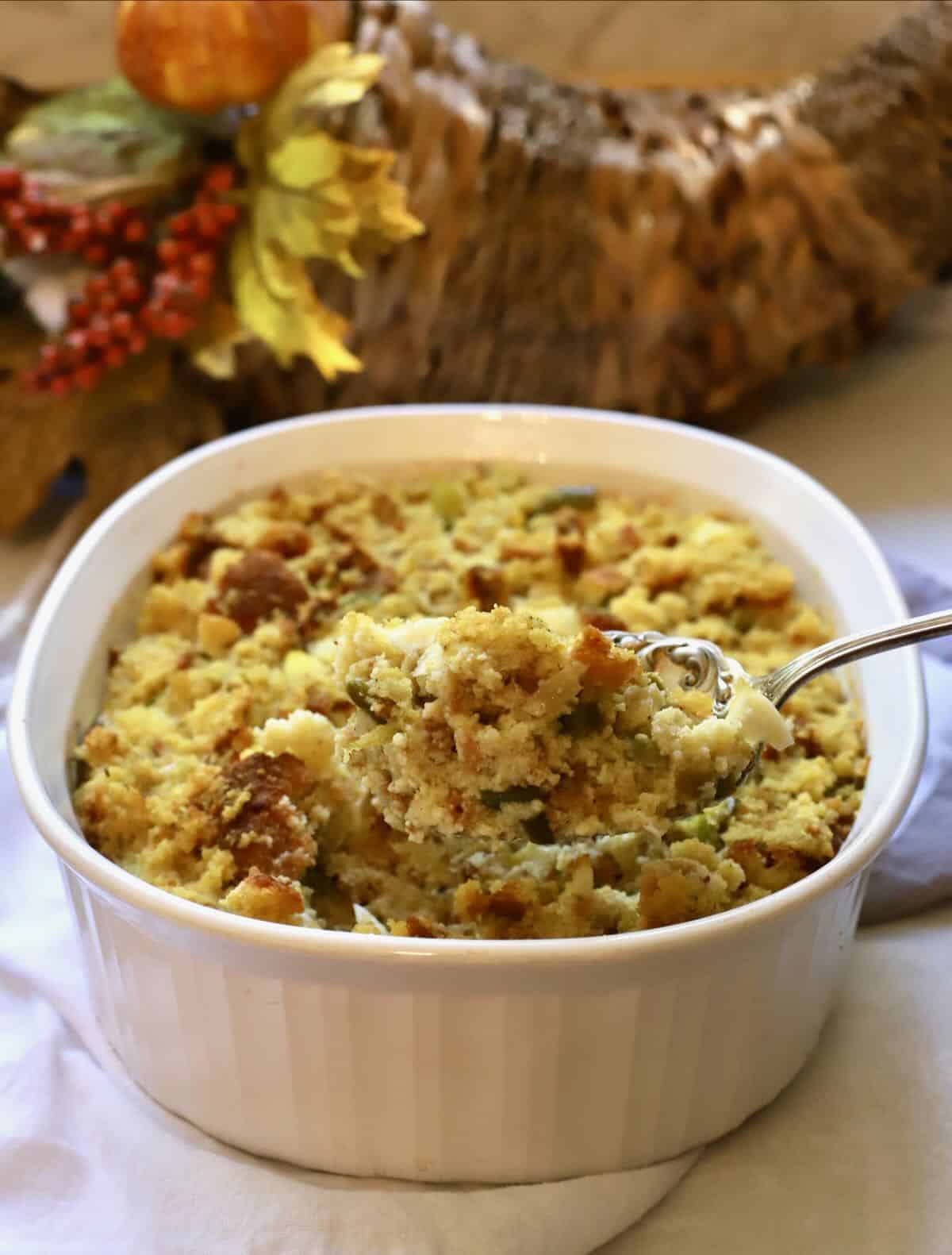 A white serving bowl full of cornbread dressing with a cornucopia in the background.