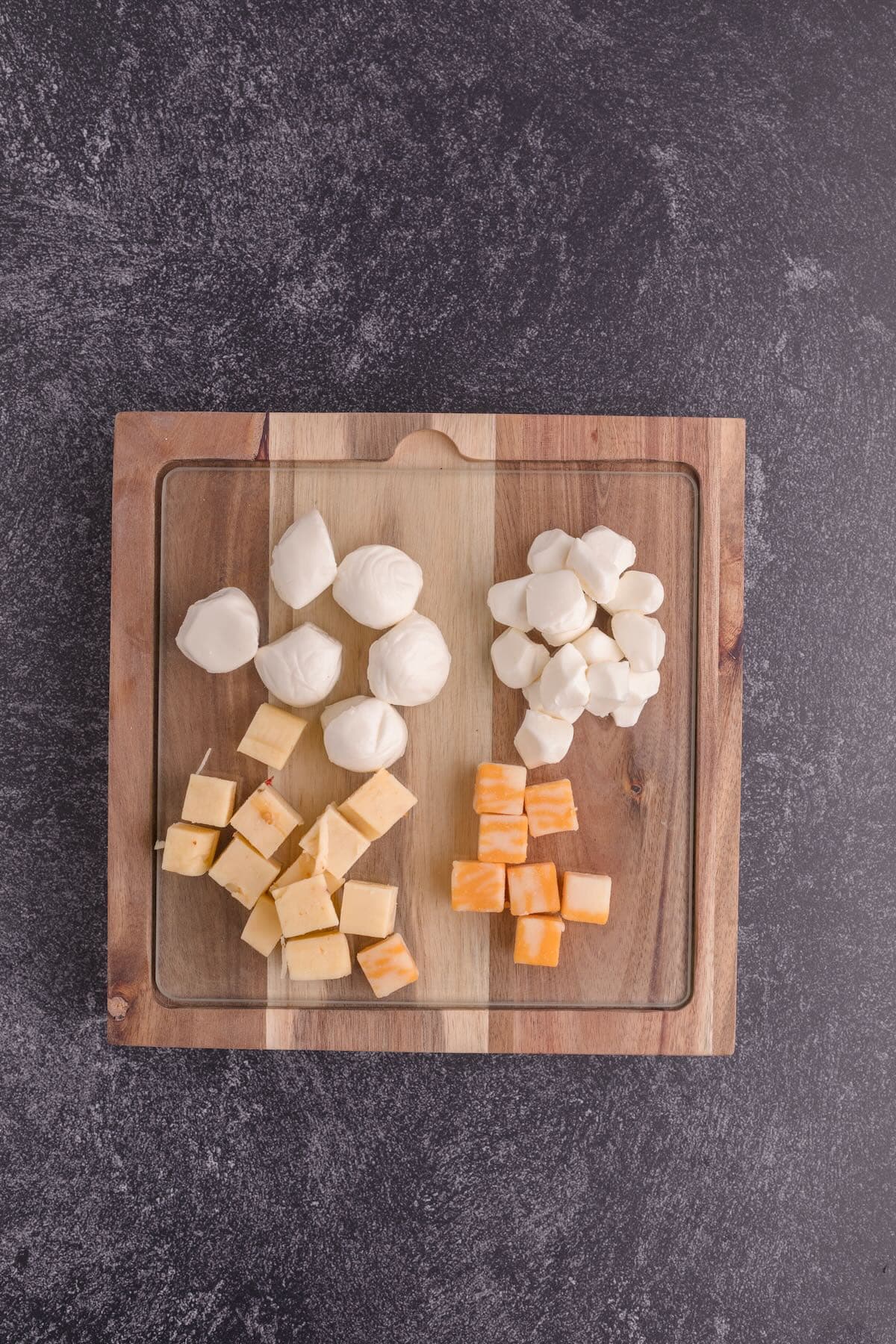 A small wooden cutting board with mozzarella balls and cubes of cheese. 