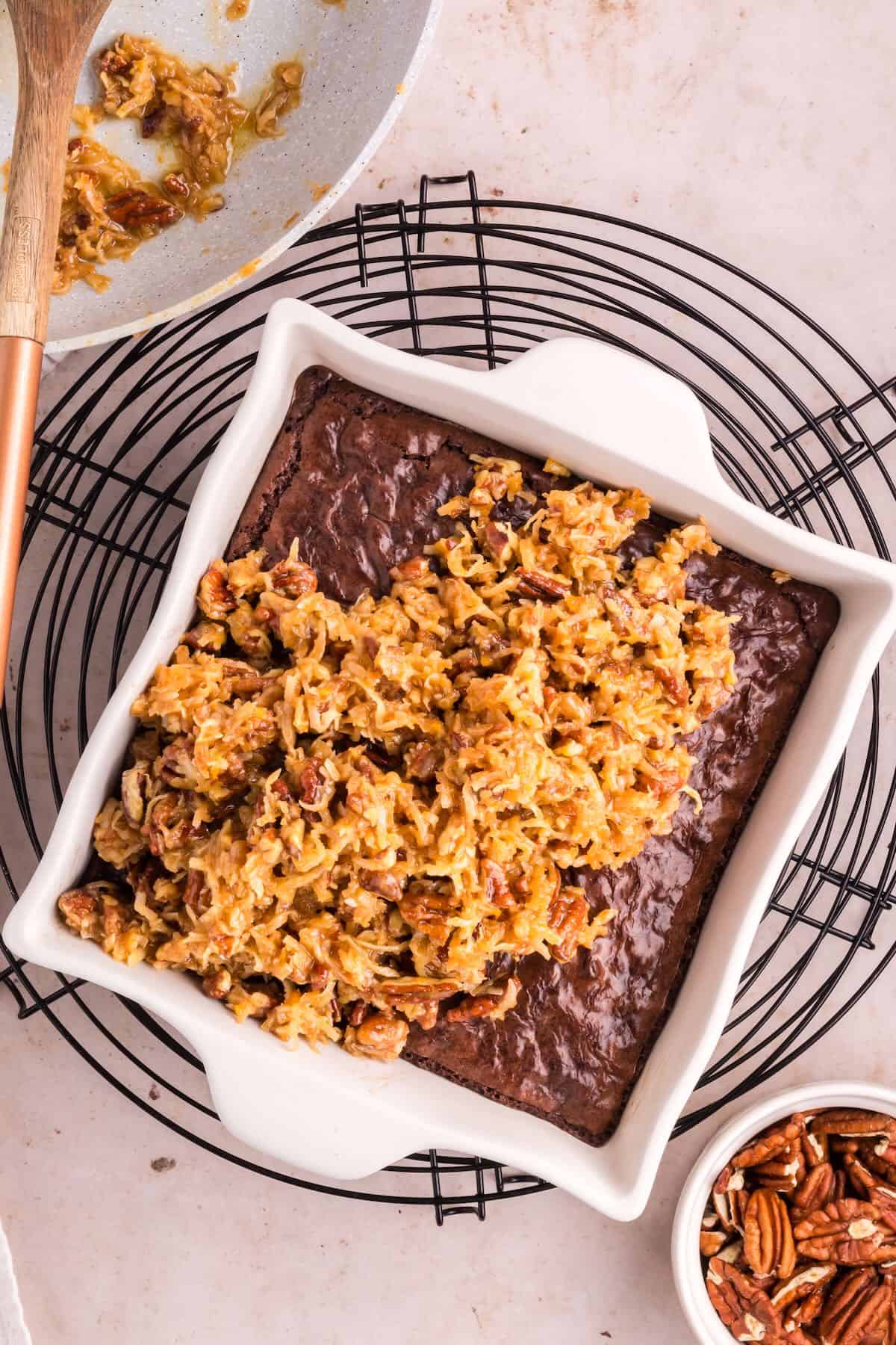 German chocolate topping is spread on top of cooked brownies in a pan. 