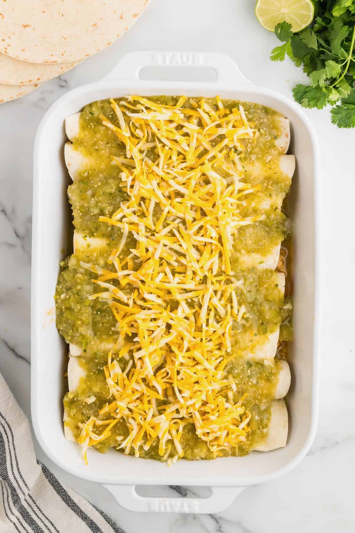 A baking dish with rolled-up enchiladas topped with green salsa verde and shredded cheese. 