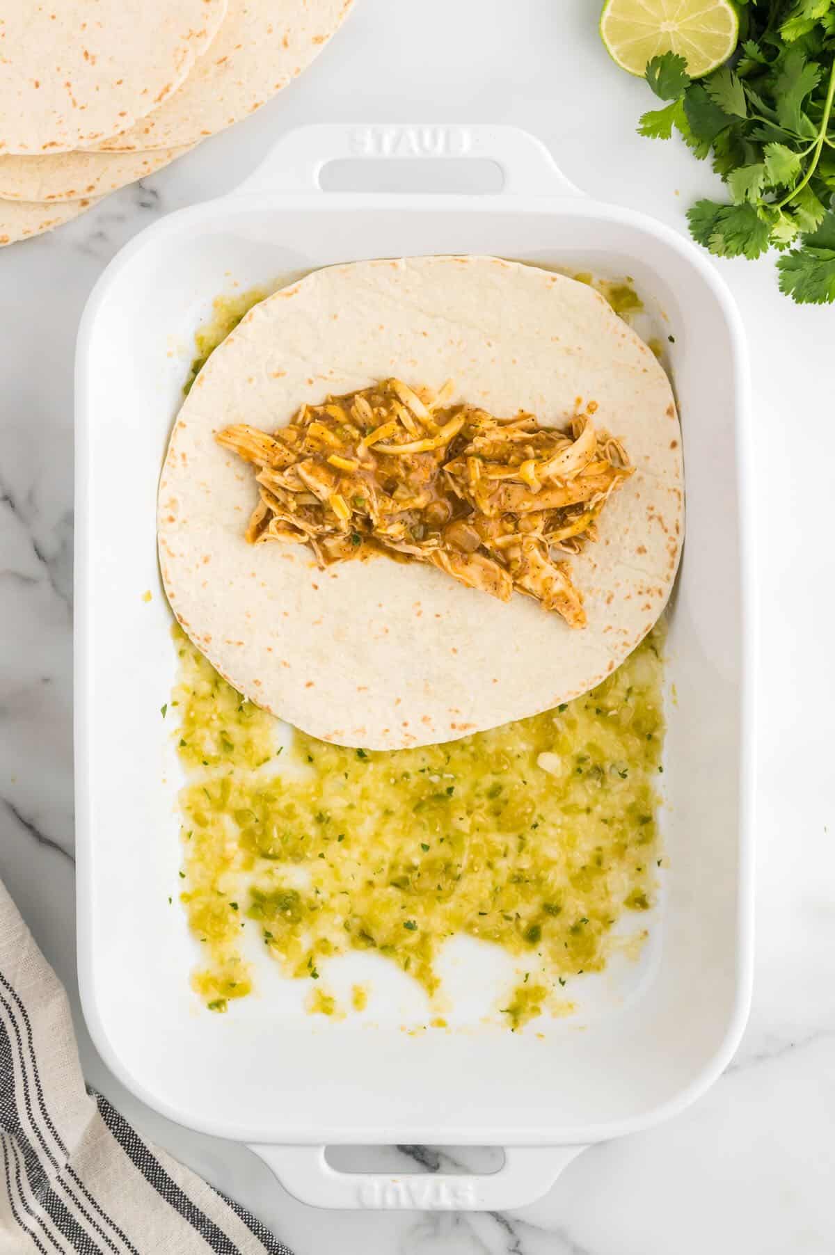 A flour tortilla with chicken enchilada filling on top of salsa verde in a baking dish. 