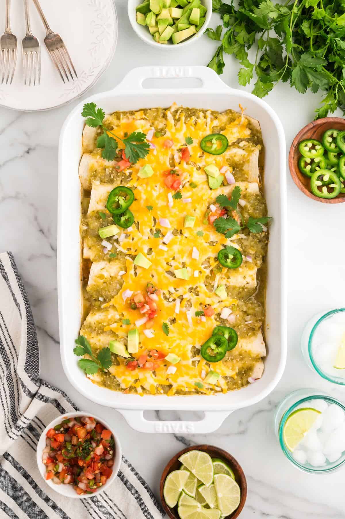 Baked green chicken enchiladas in a white baking dish topped with sliced jalapenos and tomatoes. 