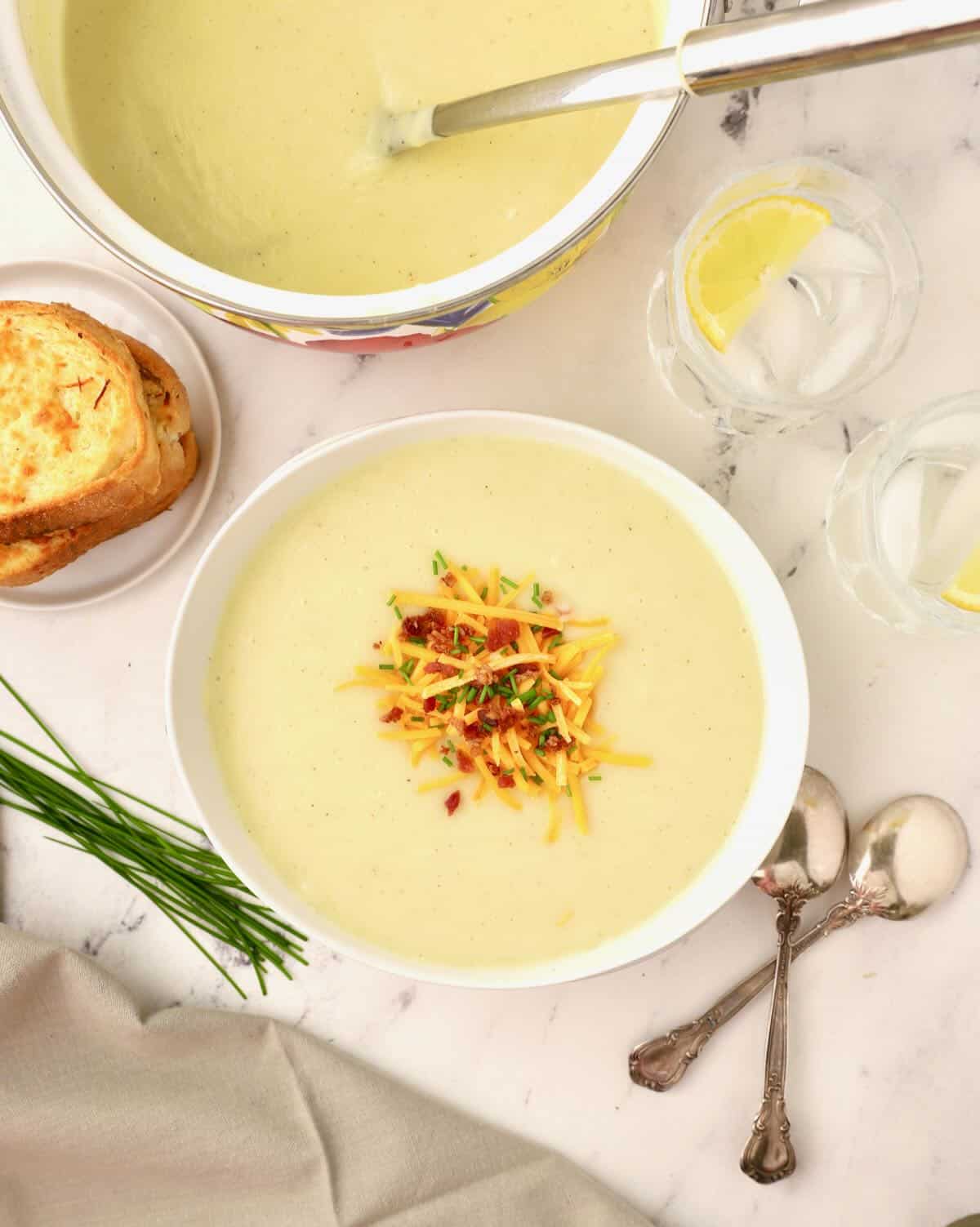 A bowl of Irish potato soup topped with cheese, bacon, and chives next to the soup pot. 