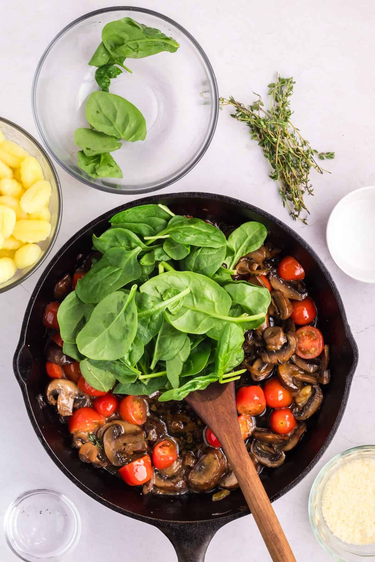 Adding spinach to a cast-iron skillet with sliced cherry tomatoes and mushrooms. 