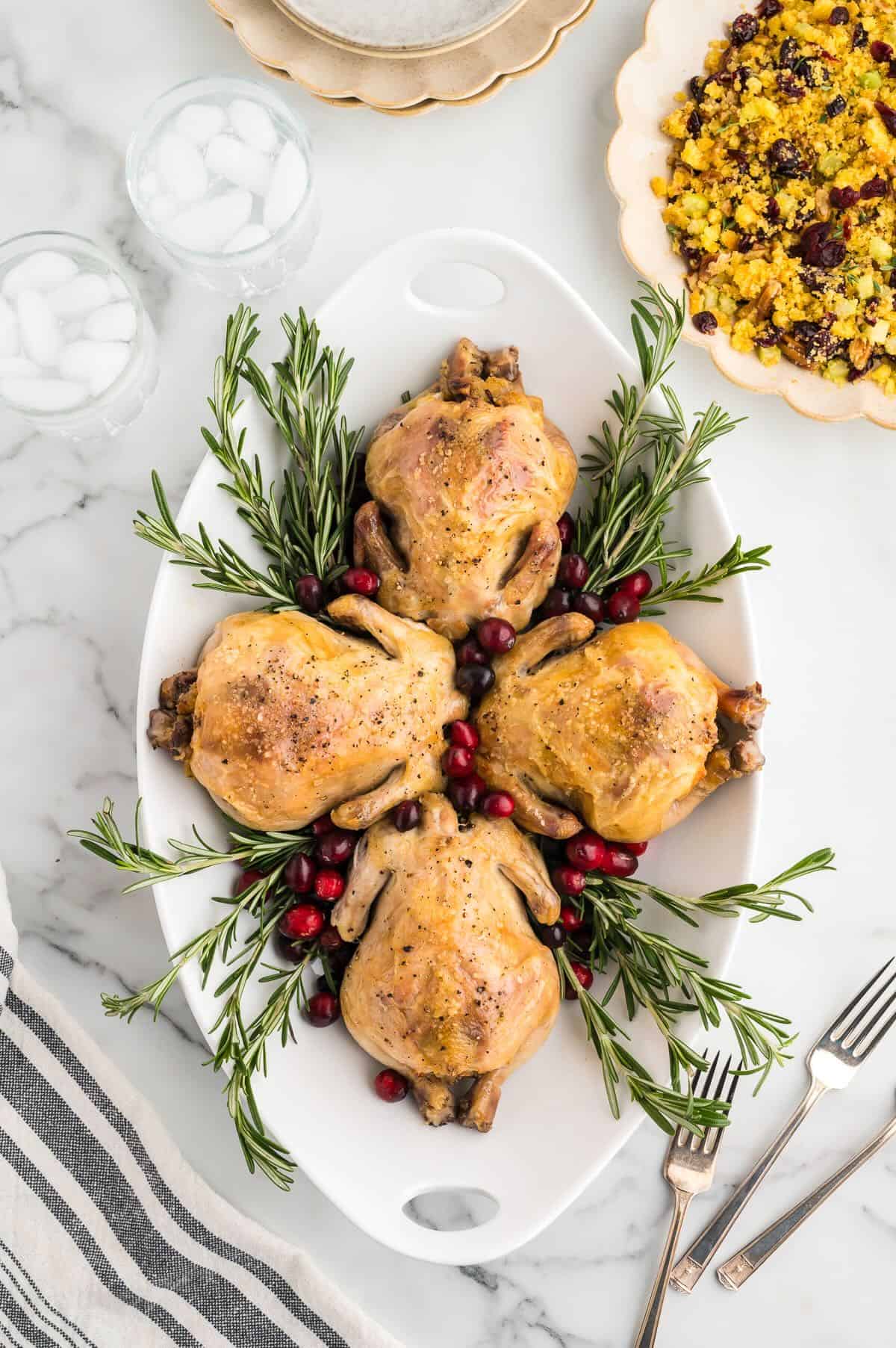 Four stuffed Cornish hens on a white platter garnished with cranberries and rosemary. 